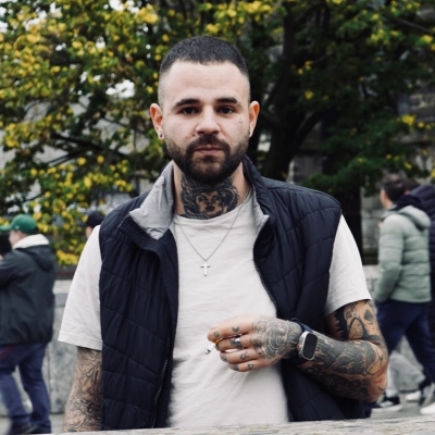 Țugui Dragoș, bearded with a short dark fade and neck and arm tattoos, wearing a white t-shirt and a black gilet, outdoors in front of autumn trees.
